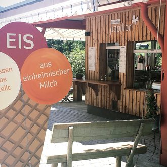 A cozy ice cream parlor with wooden paneling and a counter area. In the foreground, there is a large ice cream sign that indicates homemade ice cream. | © Papillon | Lisa Schüttler
