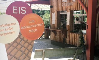 A cozy ice cream parlor with wooden paneling and a counter area. In the foreground, there is a large ice cream sign that indicates homemade ice cream. | © Papillon | Lisa Schüttler