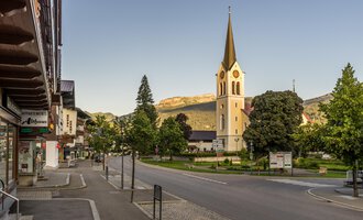 Eine ruhige Straße in einem charmanten Dorf mit einer hohen Kirche im Hintergrund. Umgeben von Bäumen und Bergen strahlt die Szene Ruhe aus. | © Kleinwalsertal Tourismus | Steffen Berschin