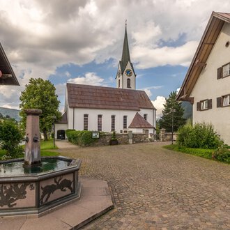 Ein malerischer Dorfplatz mit einem Brunnen im Vordergrund und traditionellen Häusern. Im Hintergrund ist eine Kirche mit einem hohen Turm und einem bewölkten Himmel zu sehen. | © Kleinwalsertal Tourismus | Steffen Berschin