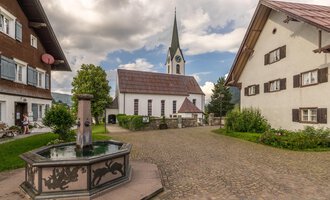 Ein malerischer Dorfplatz mit einem Brunnen im Vordergrund und traditionellen Häusern. Im Hintergrund ist eine Kirche mit einem hohen Turm und einem bewölkten Himmel zu sehen. | © Kleinwalsertal Tourismus | Steffen Berschin