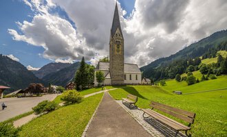 Eine malerische Kirche mit einem spitzen Turm steht in einer grünen Landschaft. Im Hintergrund sind sanfte Hügel und Wolken am Himmel zu sehen. | © Kleinwalsertal Tourismus | Steffen Berschin