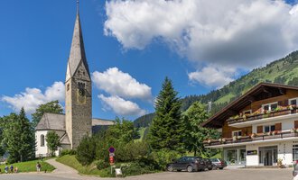 A picturesque street with a church and a traditional building. In the background, one can see green hills and a blue sky with white clouds. | © Kleinwalsertal Tourismus | Steffen Berschin