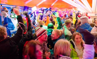 Eine lebhafte Feier mit vielen Menschen in bunten Skibekleidungen. Die Atmosphäre ist energiegeladen mit Lichtern und fröhlichen Tänzen. | © Plattform Kleinwalsertal | Michael Monschau