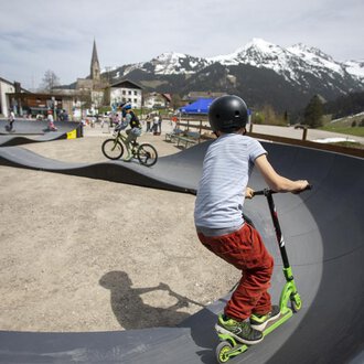 Ein Skatepark mit Kindern, die auf einem Roller und Fahrrad fahren. Im Hintergrund sind Berge und eine kleine Stadt zu sehen. | © Kleinwalsertal Tourismus | Frank Drechsel