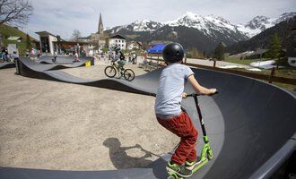 Ein Skatepark mit Kindern, die auf einem Roller und Fahrrad fahren. Im Hintergrund sind Berge und eine kleine Stadt zu sehen. | © Kleinwalsertal Tourismus | Frank Drechsel