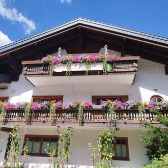 A traditional alpine house with blooming balcony plants. The sky is blue and the surroundings are green. | © Kleinwalsertal Tourismus | N. Lughammer