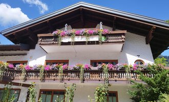 A traditional alpine house with blooming balcony plants. The sky is blue and the surroundings are green. | © Kleinwalsertal Tourismus | N. Lughammer