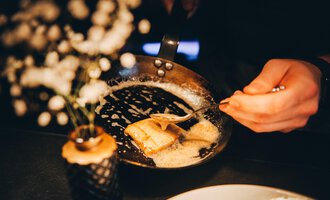 A hand serves a golden-brown dish from a pan. In the background, there is a small flower vase.