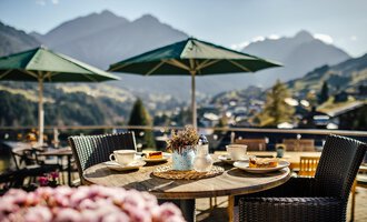 A beautiful table outdoors with dishes and pastries, surrounded by a magnificent mountain landscape. Umbrellas provide shade for a cozy atmosphere.