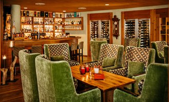 A cozy lobby and bar with green armchairs and a wooden table. In the background, there are shelves with spirits such as rum and whiskey bottles.
