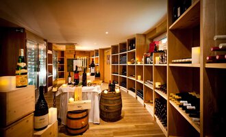 An elegant wine cellar with wooden shelves full of wine bottles. In the middle stands a table with candles and decorative elements. | © Hotel Birkenhöhe | Fotograf Krug