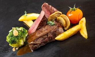 A juicy steak with roasting aromas, served with French fries, broccoli, and a tomato. The dish is accompanied by herbs and mushrooms. | © Hotel Alte Krone