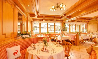 A cozy restaurant with wooden walls and elegant tablecloths. The atmosphere is enlivened by natural light sources and decorative plants. | © Aparthotel Kleinwalsertal Haller GmbH & Co KG