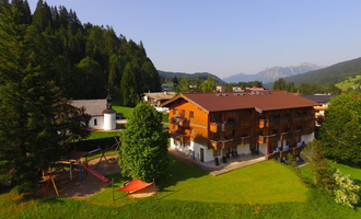 A cozy wooden house is situated in a green landscape, surrounded by mountains and trees. In the foreground, there is a playground for children. | © Hotel Bellevue | Werbewind GmbH