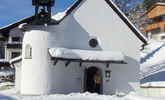 A small white chapel in the snow with a bell tower. In the background, snow-covered trees and a wooden building are visible. | © Hotel Bellevue | I. Malouvier