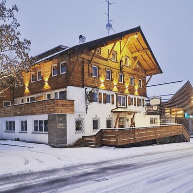 A charming wooden house in the snow, illuminated by warm light. The surroundings are wintry and inviting. | © Hotel Neue Krone GmbH | Werbewind GmbH