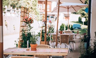 A cozy outdoor area of a café with tables and chairs under sun umbrellas. Surrounded by plants and a welcoming atmosphere. | © Hotel Neue Krone GmbH | Werbewind GmbH