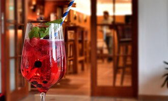 A refreshing drink in a glass with a blue straw is on a table. In the background, a cozy bar can be seen. | © Hotel Neue Krone GmbH | Werbewind GmbH