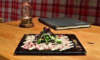 A beautifully arranged plate with fine meat and fresh herbs. In the background, a lamp and a cookbook can be seen. | © Hotel Neue Krone GmbH | Werbewind GmbH