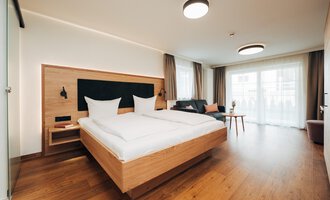 A modern hotel room with a large double bed and wooden floors. Large windows let in plenty of daylight.