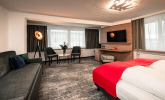 A modern hotel room with a red bed and a cozy seating area. Bright windows and stylish furniture create an inviting atmosphere.