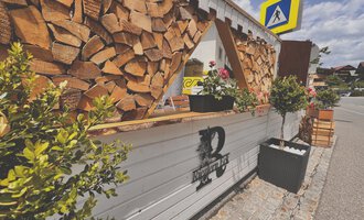 A cozy outdoor area with a wooden counter and decorative plants. In the background, wood piles and a street are visible. | © Riezlern Eck | Sead Sulejmani