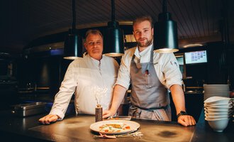 Two chefs stand in a modern kitchen behind a plate of elegantly arranged food. The atmosphere is inviting and creative, with soft lighting. | © Hotel Birkenhöhe | Stefan Klauser
