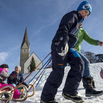 Kinder ziehen Schlitten im Schnee, während Erwachsene in warmen Kleidern sie begleiten. Im Hintergrund ist eine Kirche und der blaue Himmel zu sehen. | © Kleinwalsertal Tourismus | Dominik Berchtold