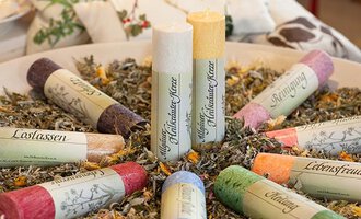 A selection of colorful candles is placed on a bed of dried herbs. Each candle is labeled with different themes. | © Rohn - Kinder, Trends und mehr|Allg. Kräuterkerzen