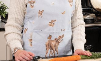 A person is wearing a stylish apron with animal motifs and is standing in a kitchen. In front of her, fresh vegetables are lying on a cutting board.