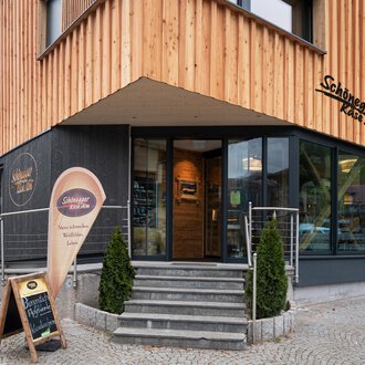 A modern restaurant with a wooden facade and large windows. The entrance area is designed to be welcoming, featuring a sign for the restaurant. | © Schönegger Käse-Alm
