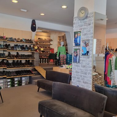 A shoe store with numerous shelves full of shoes. In the center, there are comfortable sofas, and clothing is on display. | © Schuhhaus Schuster | Marietta Diehl