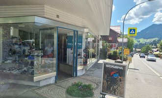 A store on a street with windows displaying shoes. In the background, mountains and a blue sky can be seen. | © Schuhhaus Schuster | Marietta Diehl