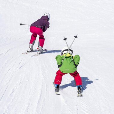 Zwei Kinder beim Skifahren auf einer schneebedeckten Piste. Das eine Kind trägt einen grünen Anzug, das andere einen lila Anzug. | © Kleinwalsertal Tourismus | Dominik Berchtold