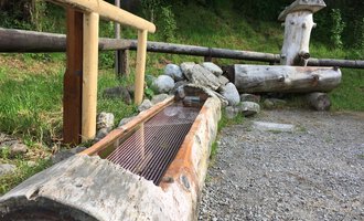 A wooden fountain made of tree trunks stands next to a stone and grass area. Behind it, more wooden structures can be seen, surrounded by trees. | © Kleinwalsertal Tourismus