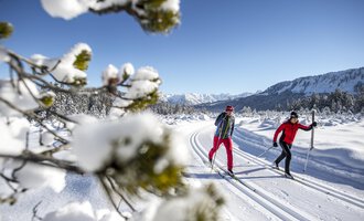 Zwei Personen skifahren auf einer verschneiten Strecke. Im Hintergrund sind schneebedeckte Berge und ein klarer blauer Himmel zu sehen. | © Kleinwalsertal Tourismus | Dominik Berchtold