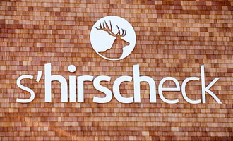 A logo with the name "s'hirschcheck" is placed on a wooden surface. The design features a stylized antler in a round cutout. | © Kleinwalsertal Tourismus | Frank Drechsel