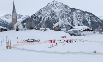 Eine verschneite Landschaft mit einem Dorf und einer Kirche im Vordergrund. Im Hintergrund ragen steile Berge empor, während eine Gruppe von Menschen in roten Jacken aktiv ist. | © Skischule Mittelberg | Karl Schuster