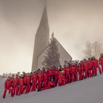 Eine Gruppe von Menschen in roten Skianzügen steht im Schnee vor einer Kirche. Die Landschaft ist winterlich und von Nebel umgeben. | © Skischule Mittelberg | Karl Schuster