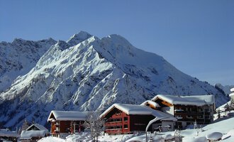 Ein malerisches Bergdorf im Schnee mit traditionellen Holzhäusern. Im Hintergrund erheben sich majestätische, schneebedeckte Gipfel. | © IFA Hotel Alpenrose