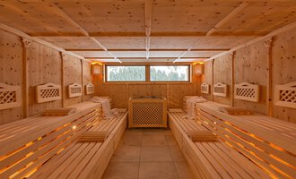 A cozy sauna room made of light wood with benches and indirect lighting. The windows provide a view of the greenery. | © Aparthotel Kleinwalsertal Haller GmbH & Co KG