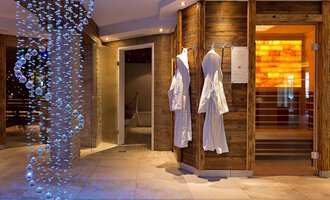 A modern wellness interior with soft lighting. On the left, white bathrobes hang, and an impressive water installation gives the room a relaxing atmosphere. | © Aparthotel Kleinwalsertal Haller GmbH & Co KG