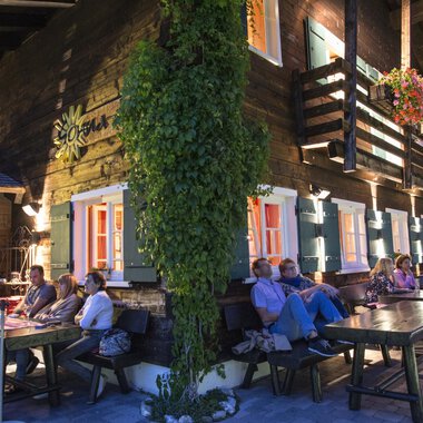 Ein gemütliches Holzhaus mit bunten Fenstern und Pflanzen. Darauf sitzen mehrere Personen an langen Tischen und genießen die Abendstimmung. | © Sonna Alp | Wolfram Riezler