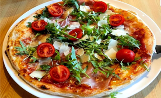 A delicious pizza with ham, cherry tomatoes, and fresh arugula. The topping is garnished with shavings of Parmesan.
