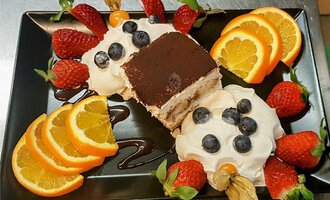 A delicious dessert on a black plate, consisting of tiramisu and fluffy cream. It is surrounded by fresh strawberries, blueberries, and orange slices.