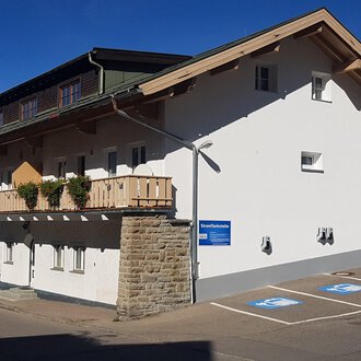 Ein modernes Gebäude mit einem Balkon und einem Parkplatz. Die Fassade ist hell und freundlich, umgeben von Bergen. | © Kleinwalsertal Tourismus