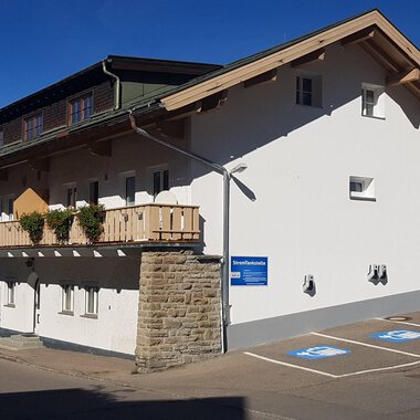 Ein modernes Gebäude mit einem Balkon und einem Parkplatz. Die Fassade ist hell und freundlich, umgeben von Bergen. | © Kleinwalsertal Tourismus