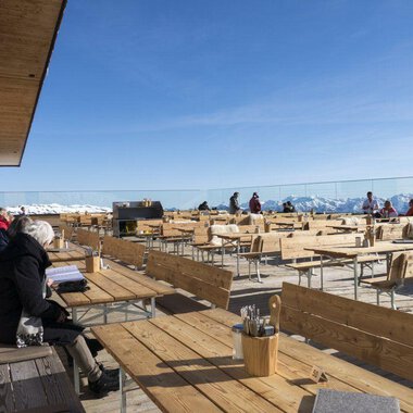 Ein idyllisches Restaurant auf einer Bergterrasse mit einer Vielzahl von Holztischen und Stühlen. Im Hintergrund sind schneebedeckte Berge und ein klarer Himmel zu sehen. | © OBERSTDORF · KLEINWALSERTAL BERGBAHNEN