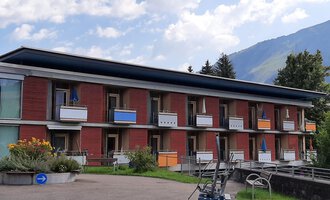 A modern building with several balconies in a green environment. In the background, mountains and a blue sky can be seen. | © Kleinwalsertal Tourismus | Veronika Senn